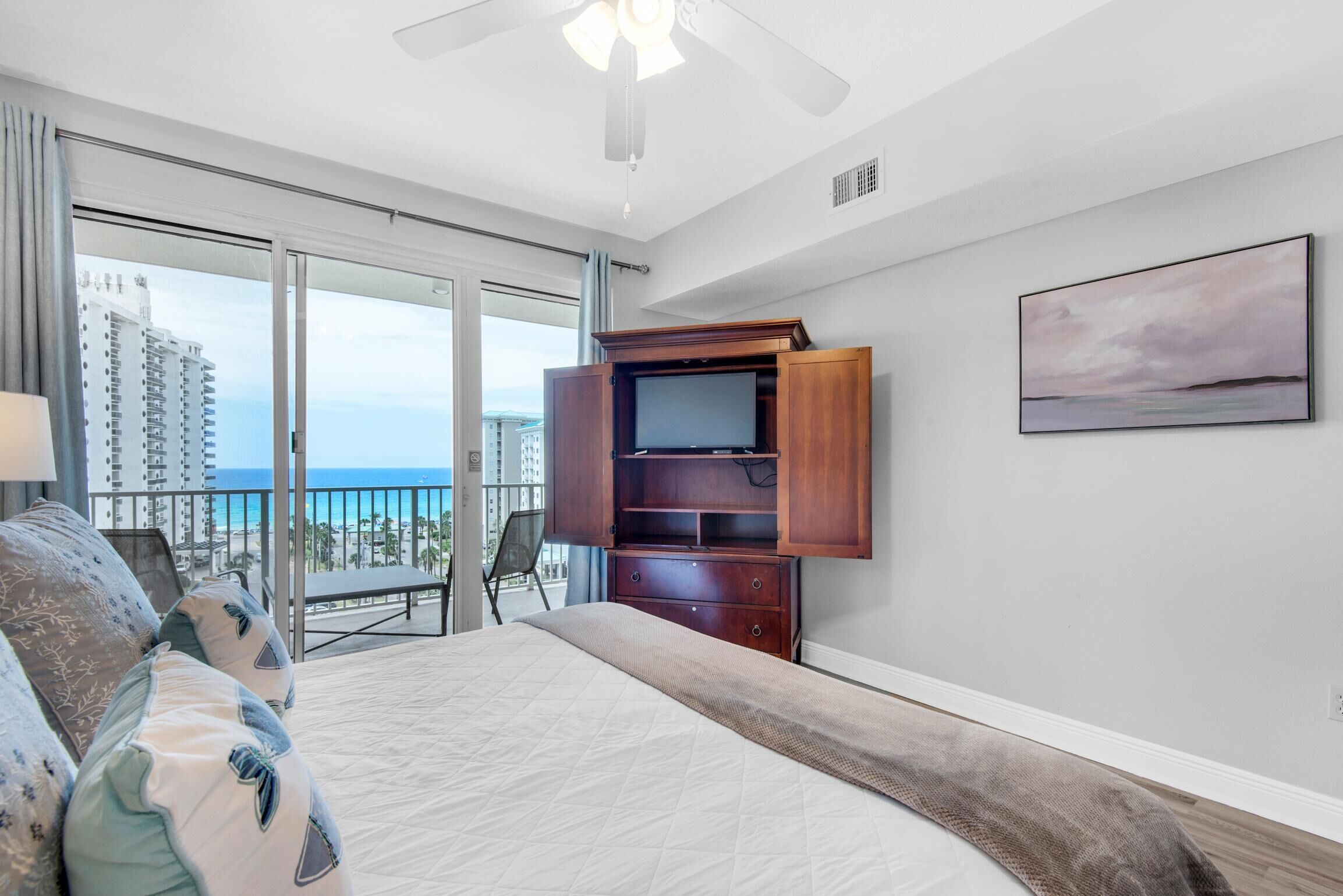 Ein Schlafzimmer mit einem großen Bett, einer Holz-TV-Konsole, einem Balkon mit Blick auf das Meer und Gebäude sowie einem Bild an der Wand.