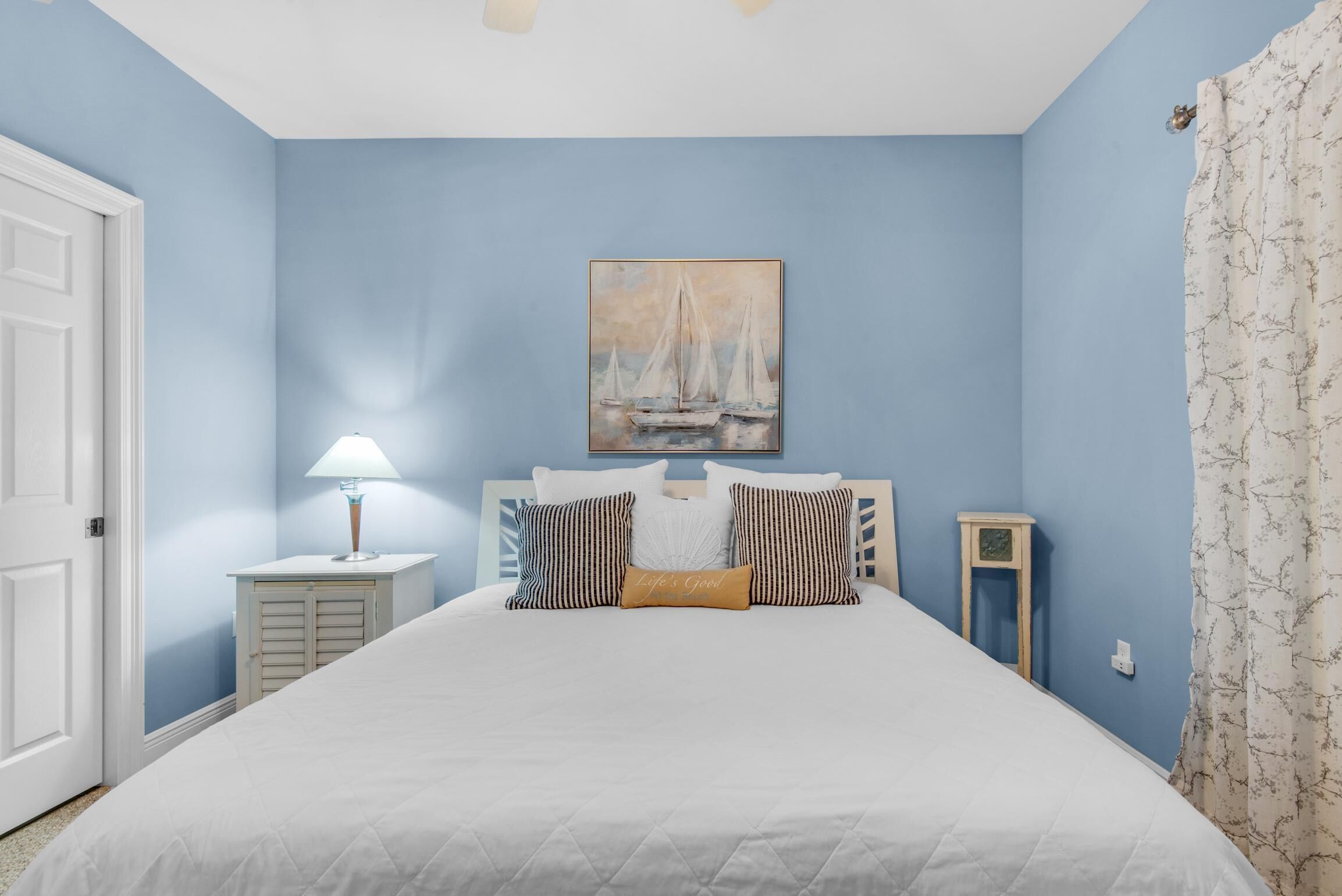 A bedroom with a bed, a painting of sailboats, a nightstand with a lamp, and a small table.