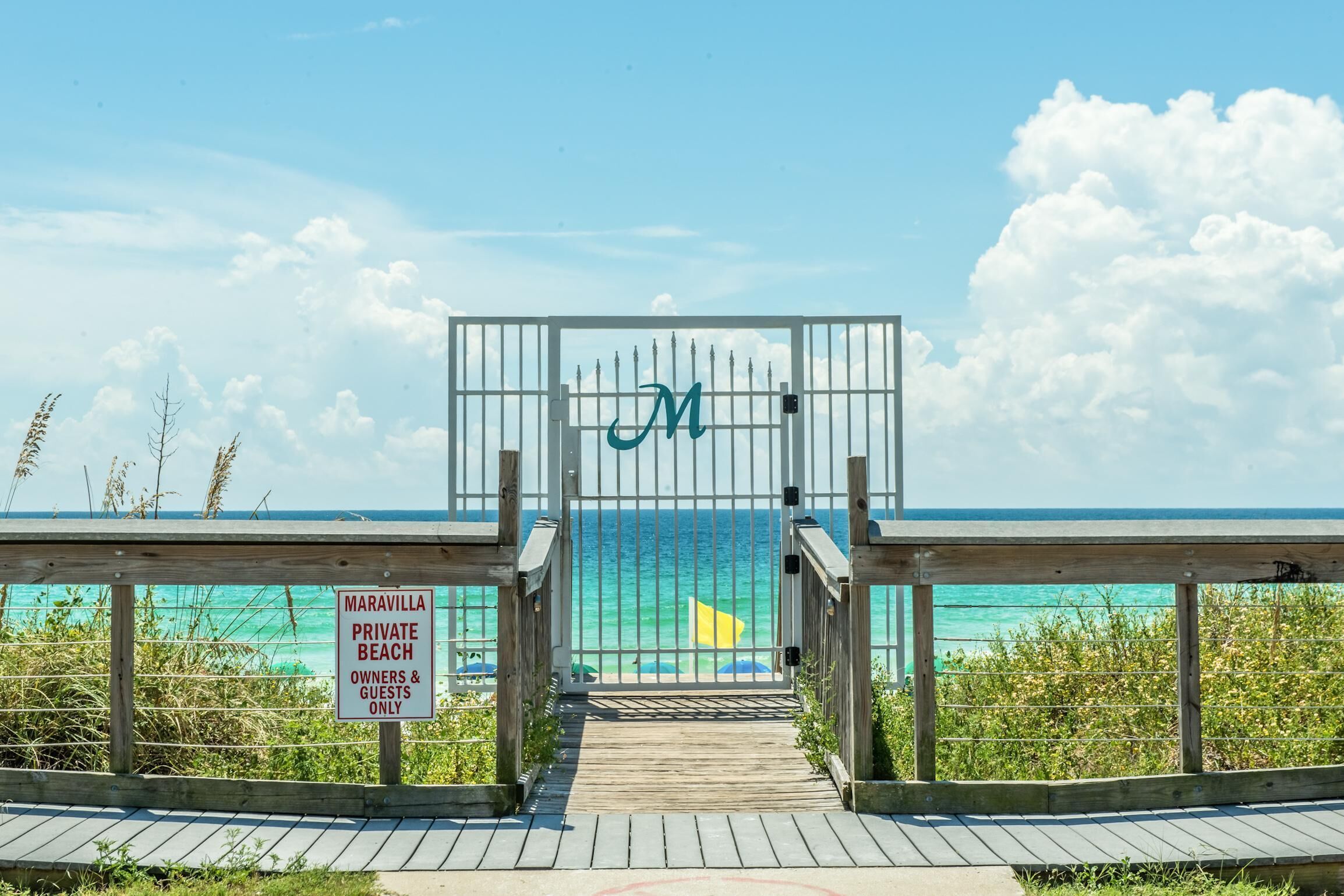 A wooden boardwalk leading to a gated entrance with a 'M' sign, labeled 'Maravilla Private Beach Owners & Guests Only'.