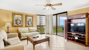 A living room with a sofa, armchair, coffee table, TV stand, ceiling fan, and large windows with a view of buildings. - Mainsail 227 (Miramar Beach)