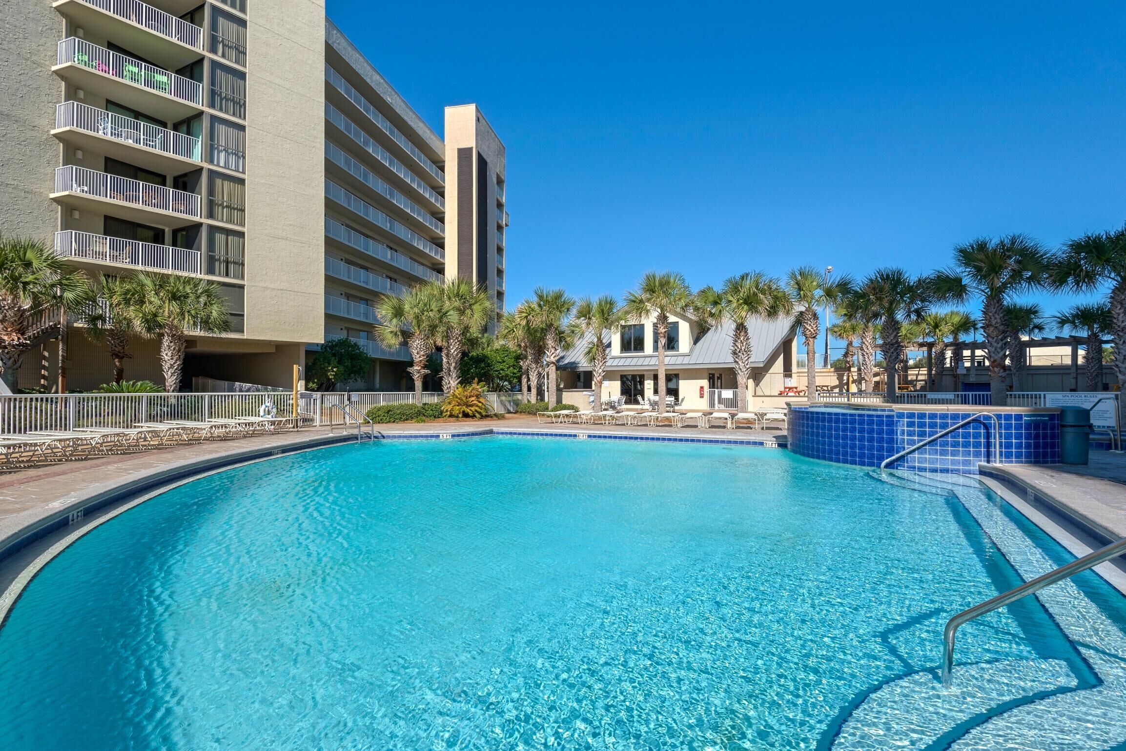 A swimming pool with a curved edge, surrounded by palm trees and a building with balconies.
