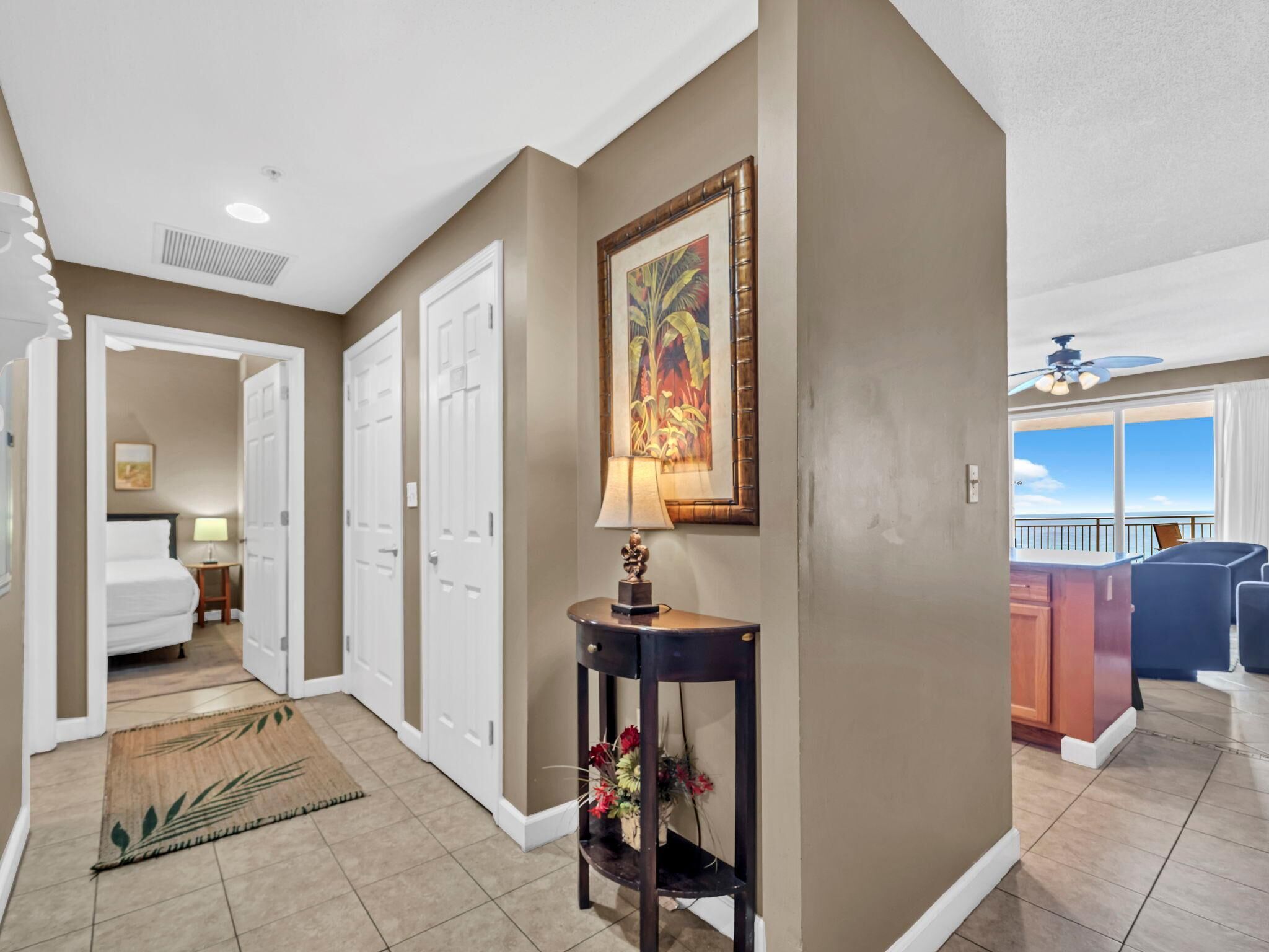 A hotel hallway with a view to a balcony, a framed painting, and a console table with flowers.