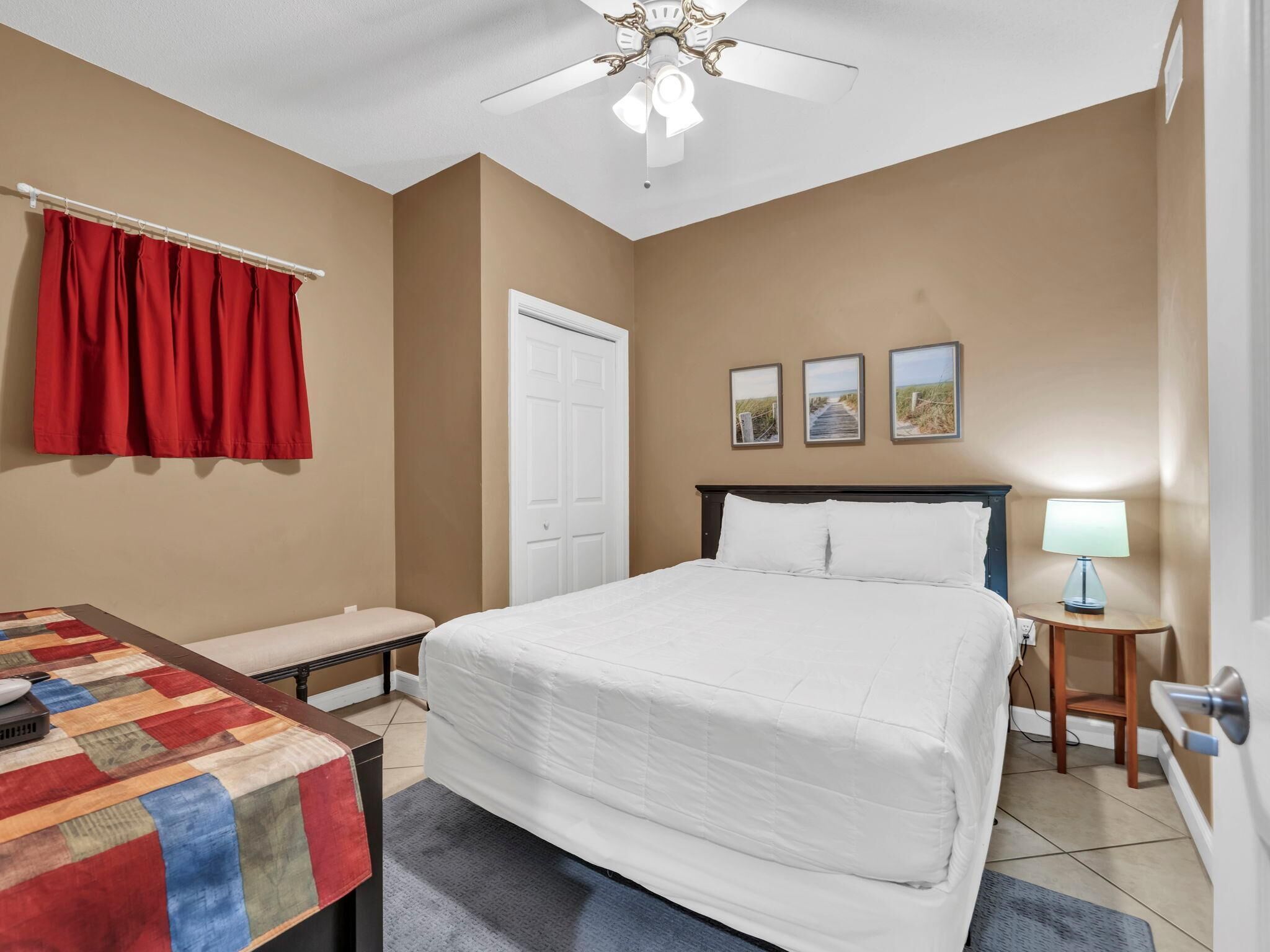 A hotel room with a bed, a desk with a chair, a red curtain, a white door, a ceiling fan, and three framed pictures on the wall.