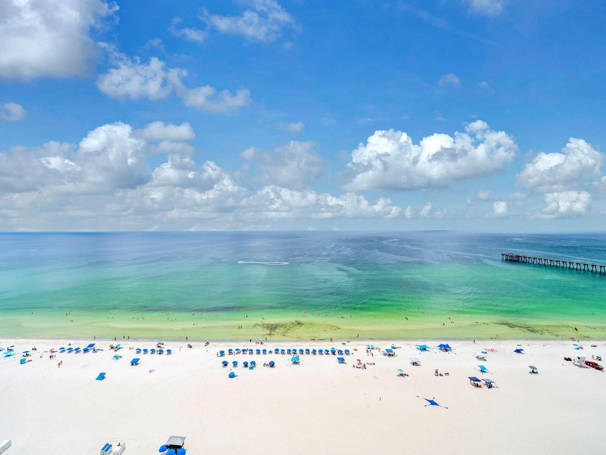 Une plage aux eaux turquoise et limpides, une jetée qui s’avance dans la mer, et de nombreux parasols bleus qui parsèment le sable.