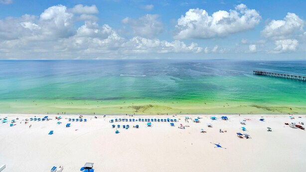 Ein Strand mit klarem türkisblauem Wasser, einer in die See ragenden Mole und zahlreichen blauen Strandsonnenschirmen.