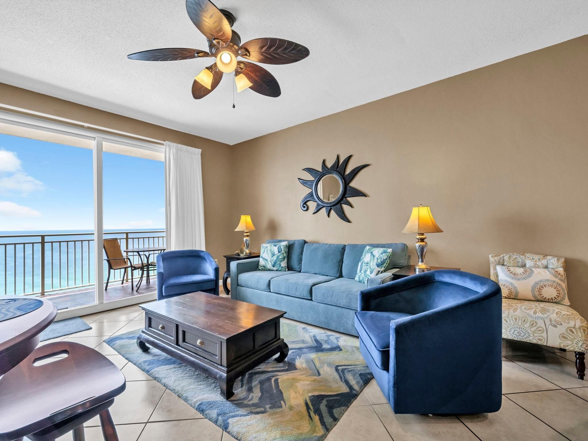 A living room with a sofa, two armchairs, a coffee table, and a ceiling fan. There is a large window with a view of the ocean.