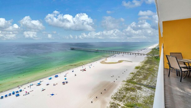 A beach view with a pier extending into the ocean, a sandy beach with umbrellas, and a balcony with chairs. - Sterling Reef 1203 (Panama City Beach)