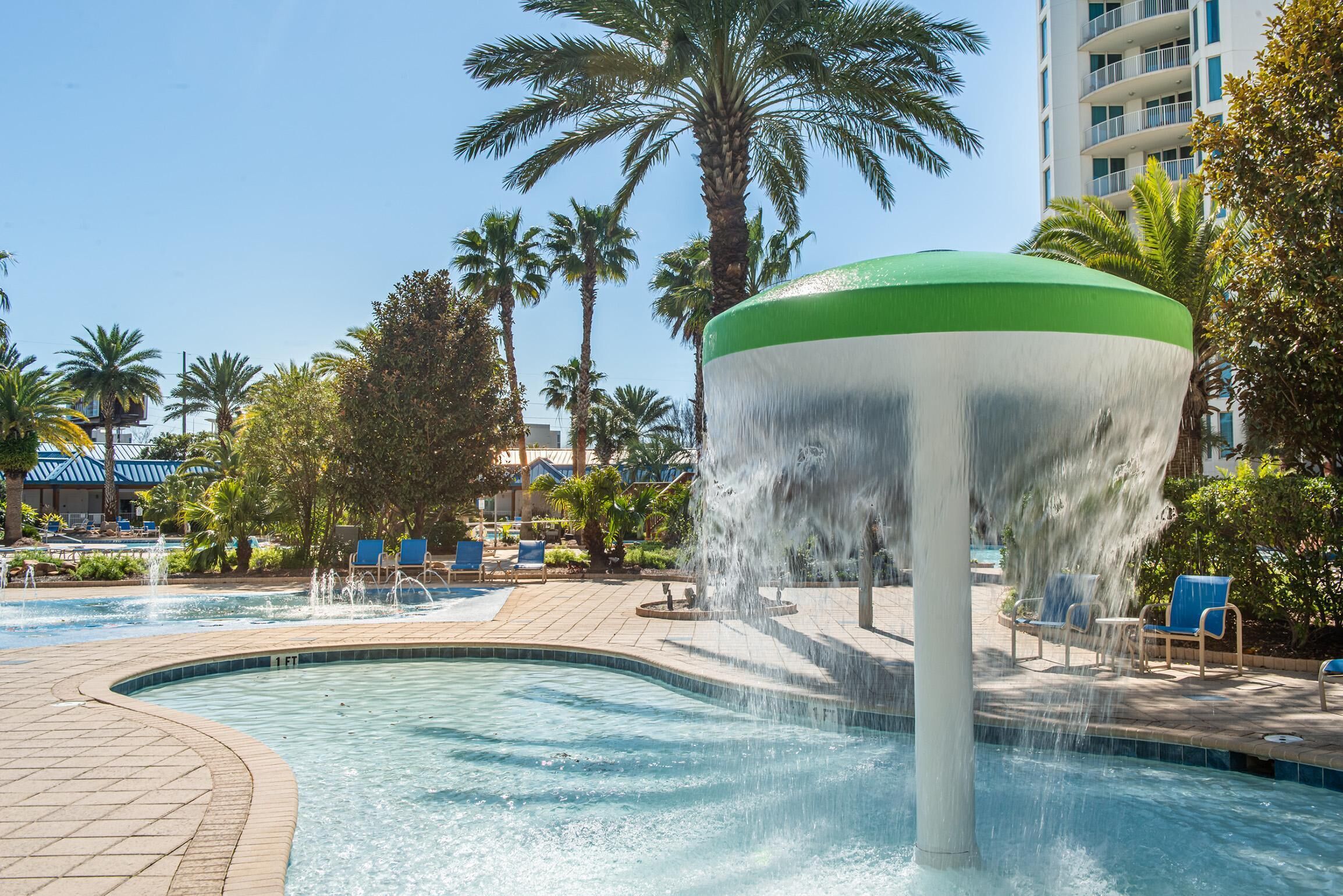 A pool area with a water fountain, palm trees, and lounge chairs.