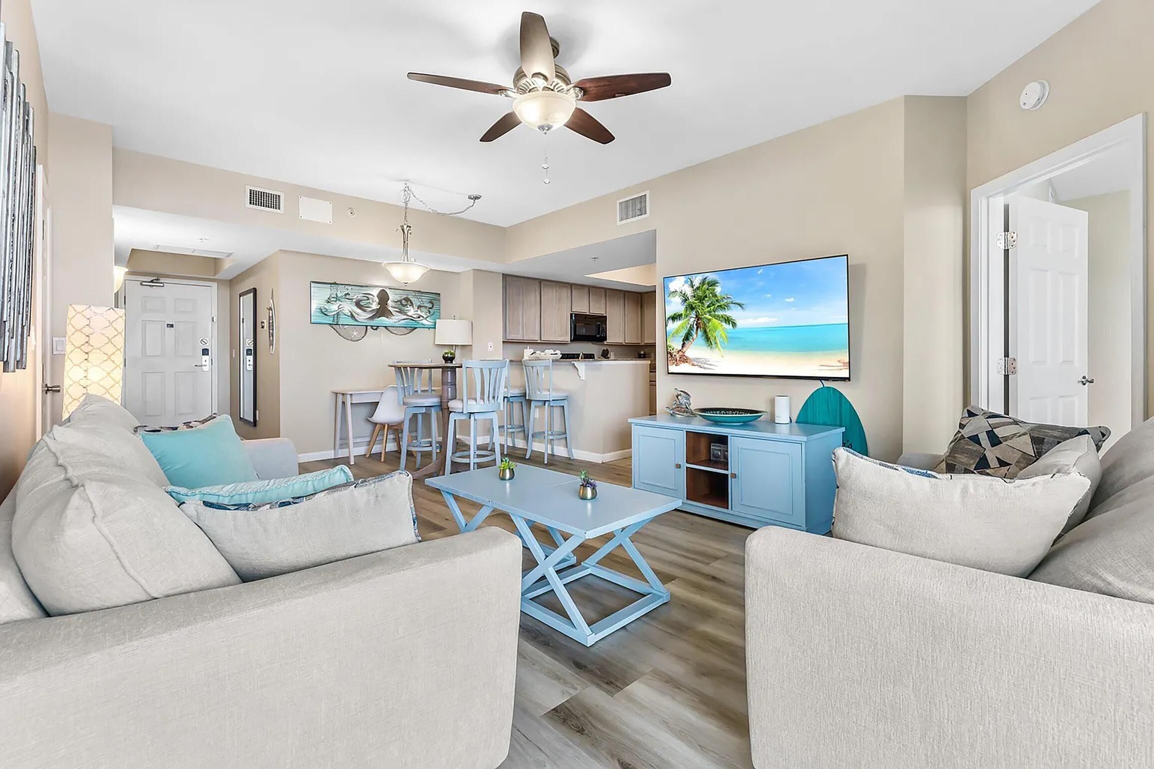 A spacious living room with a ceiling fan, a dining area with a table and chairs, a kitchen area, and a large TV displaying a beach scene.