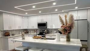 A modern kitchen with white cabinetry, a central island with bar stools, and a built-in oven. - Beach House,2 minutes walking to the beach and Restaurant The Beach House by 604 (Waianae)