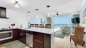A modern kitchen with a central island, dark wooden cabinets, and a stainless steel dishwasher. There is a dining area with a view of the ocean and a balcony with a chair. - Boardwalk 209 (Panama City Beach)