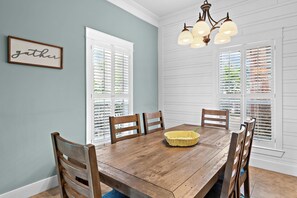Une salle à manger avec une table en bois, des chaises, un lustre et une fenêtre avec des stores.