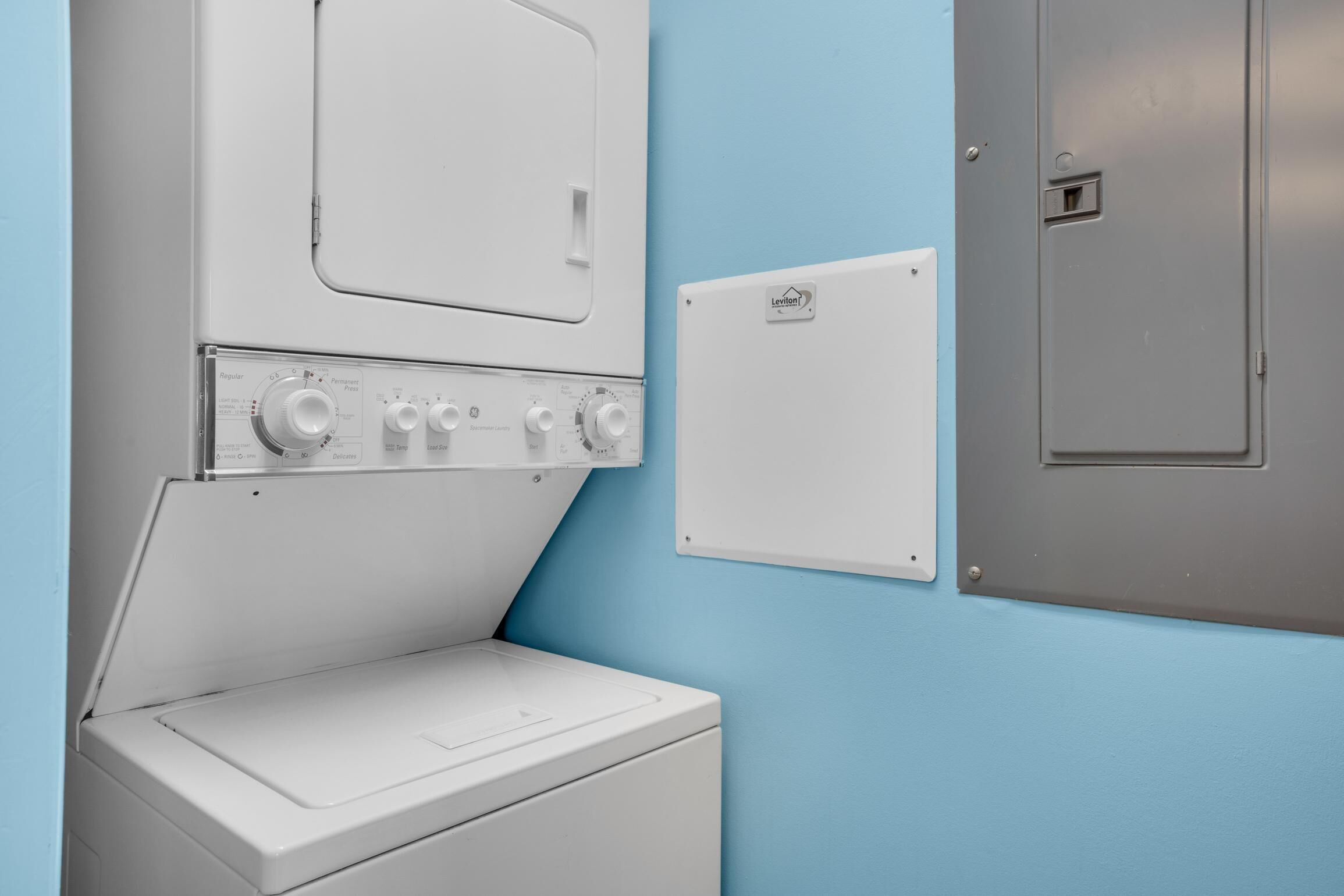 Zona de lavandería con lavadora y secadora de carga frontal, panel de control y medidor de servicios en la pared.