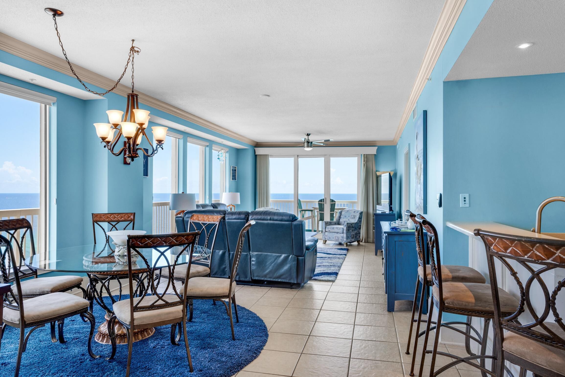 A spacious living area with a view of the ocean, featuring a chandelier, blue sofas, and a dining table with chairs.