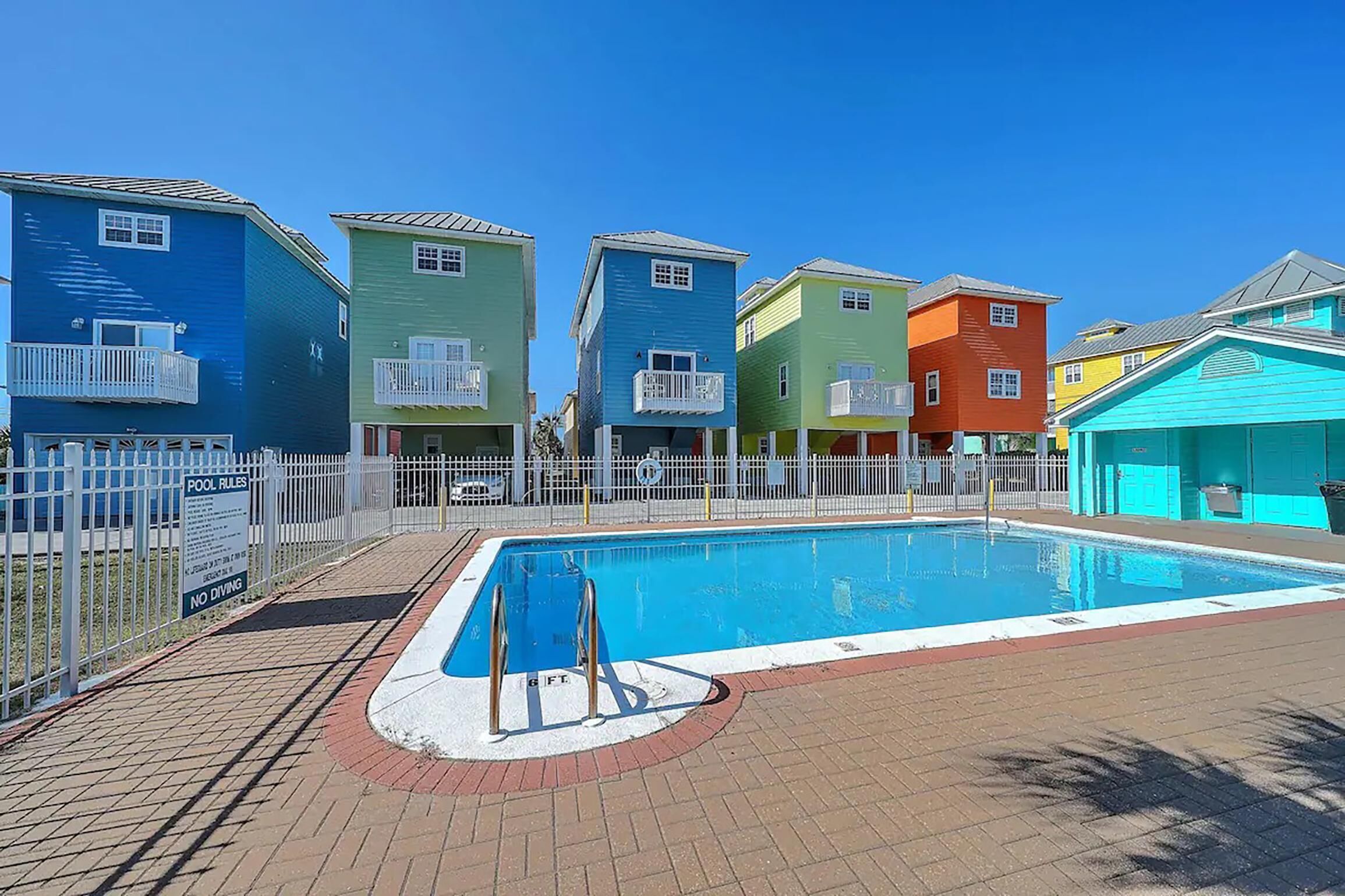 A swimming pool surrounded by colorful houses with balconies, a fenced pool area, and a clear blue sky.