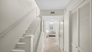 A clean, white interior hallway with a staircase, a door with louvered shutters, and a tiled floor.
