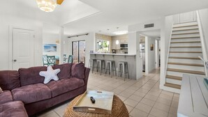 A modern living room with a sofa, a starfish pillow, and a remote control on a woven tray. A kitchen with bar stools and a staircase is visible in the background. - Seascape 51A - Beach Baby (Miramar Beach)
