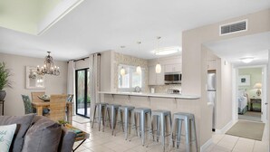 A modern kitchen with bar stools, a chandelier, and a dining area with a table and chairs. - Seascape 51A - Beach Baby (Miramar Beach)