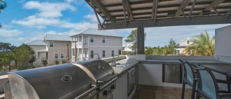 An outdoor kitchen area with a built-in grill and sink, surrounded by a covered patio with a view of neighboring houses.
