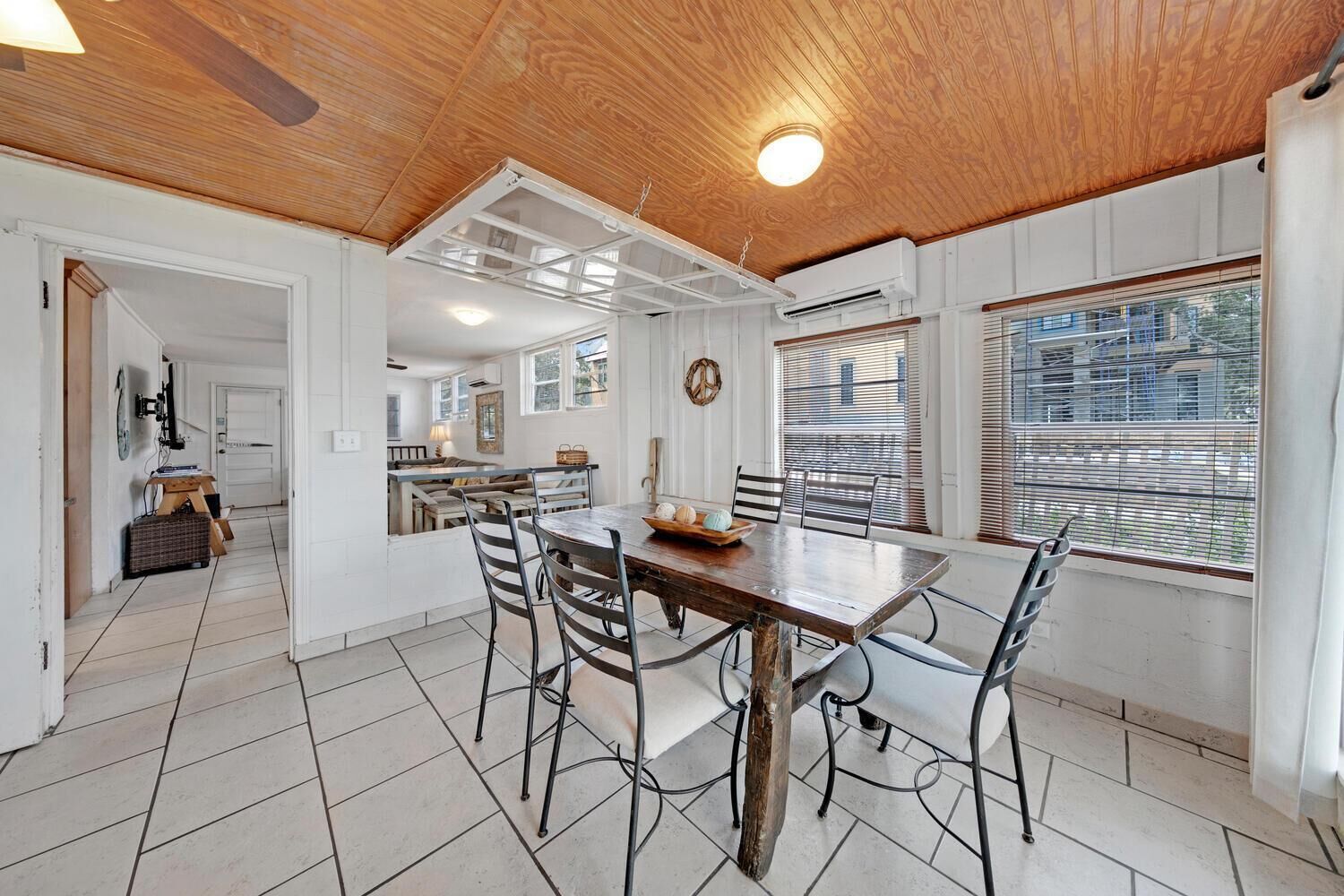 A spacious dining area with a wooden table and metal chairs, a kitchen in the background, and large windows with blinds.