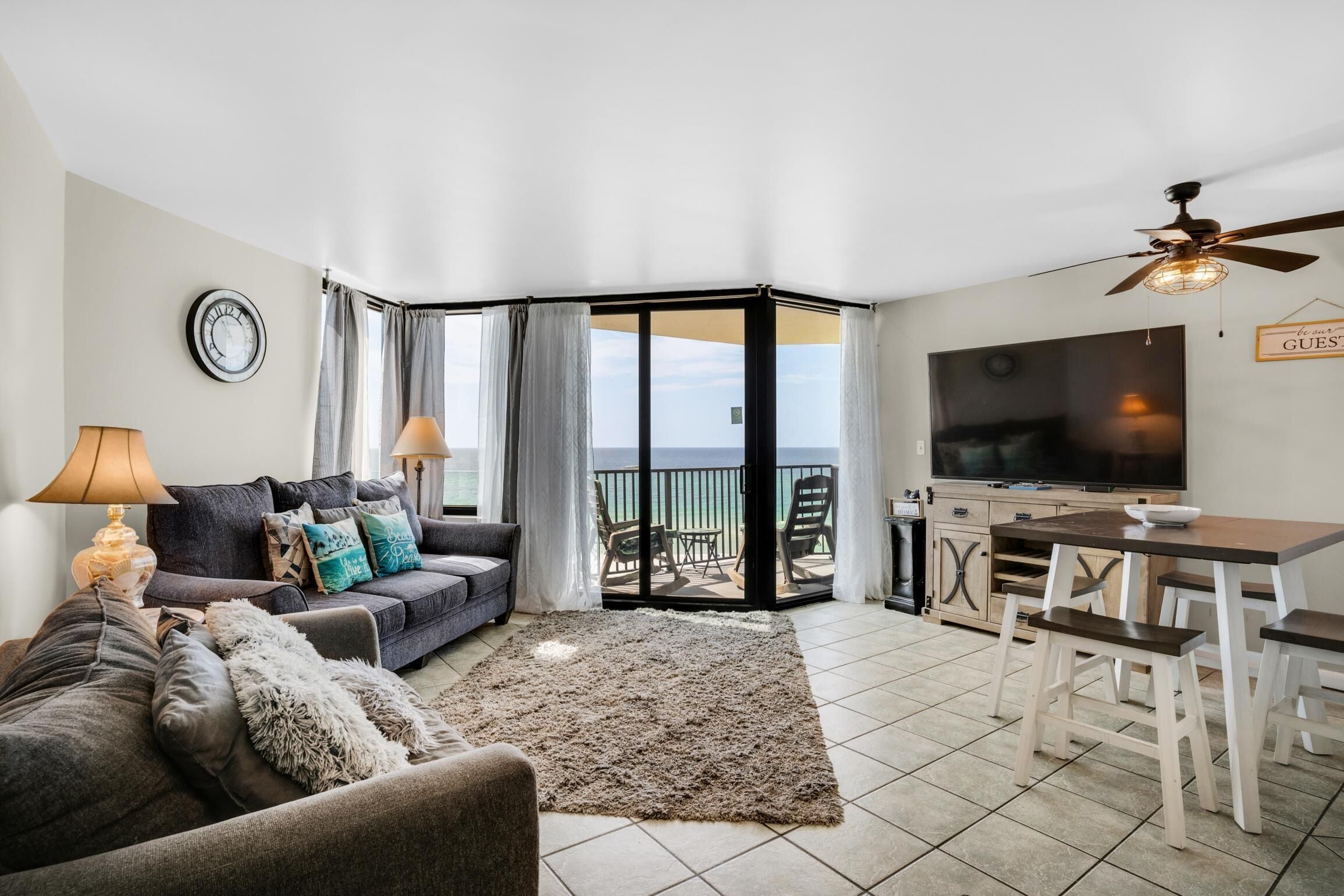 A living room with a sofa, two lamps, a TV, a dining table with chairs, and a view of the sea through a glass door.