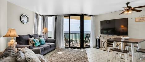 A living room with a sofa, two lamps, a TV, a dining table with chairs, and a view of the sea through a glass door.