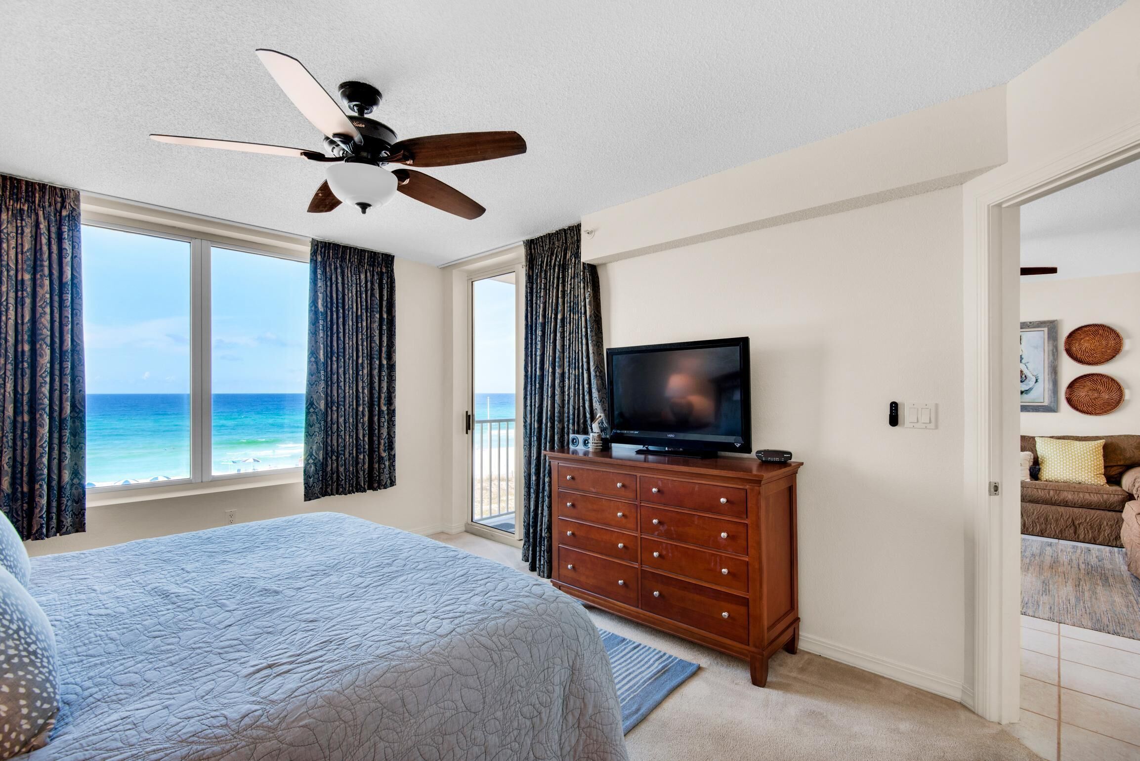 A bedroom with a bed, a television on a wooden dresser, a ceiling fan, and a view of the ocean through large windows.