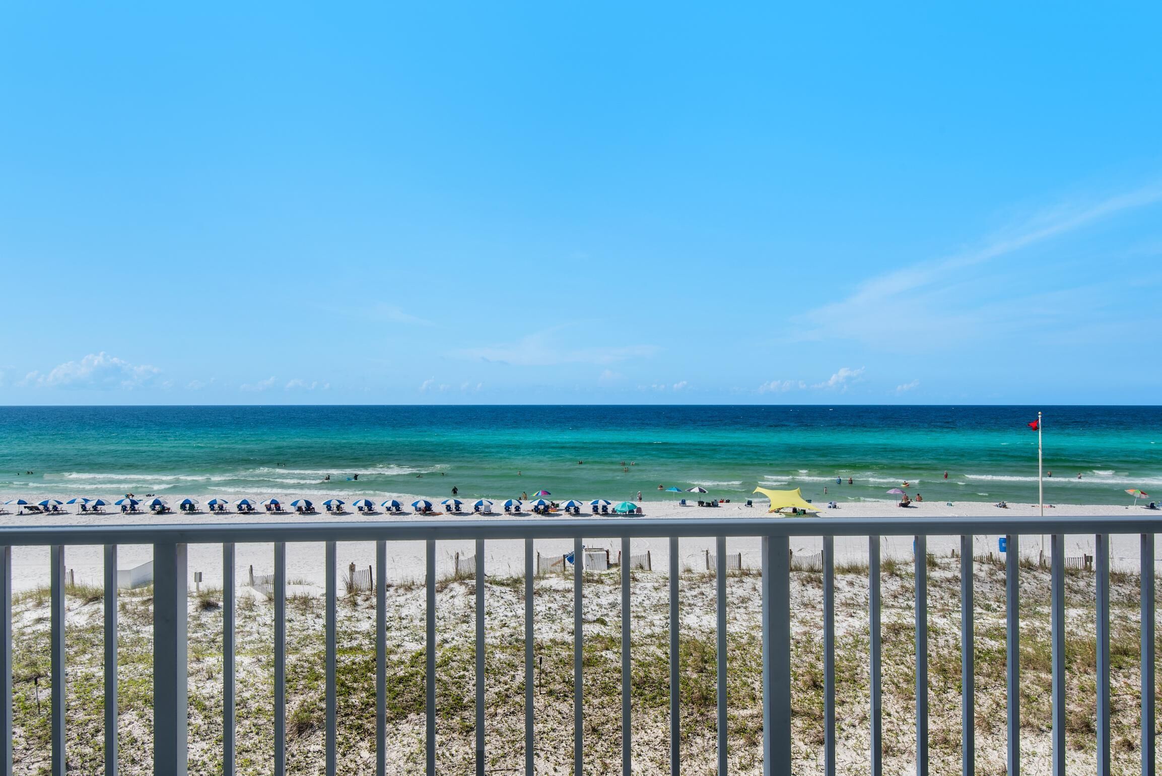 Ein Strand mit weißem Zaun, Sonnenschirmen, einer Flagge und wolkenlosem, blauem Himmel.
