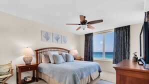 A bedroom with a bed, two bedside tables, a desk, a ceiling fan, a window with curtains, and artwork on the wall.