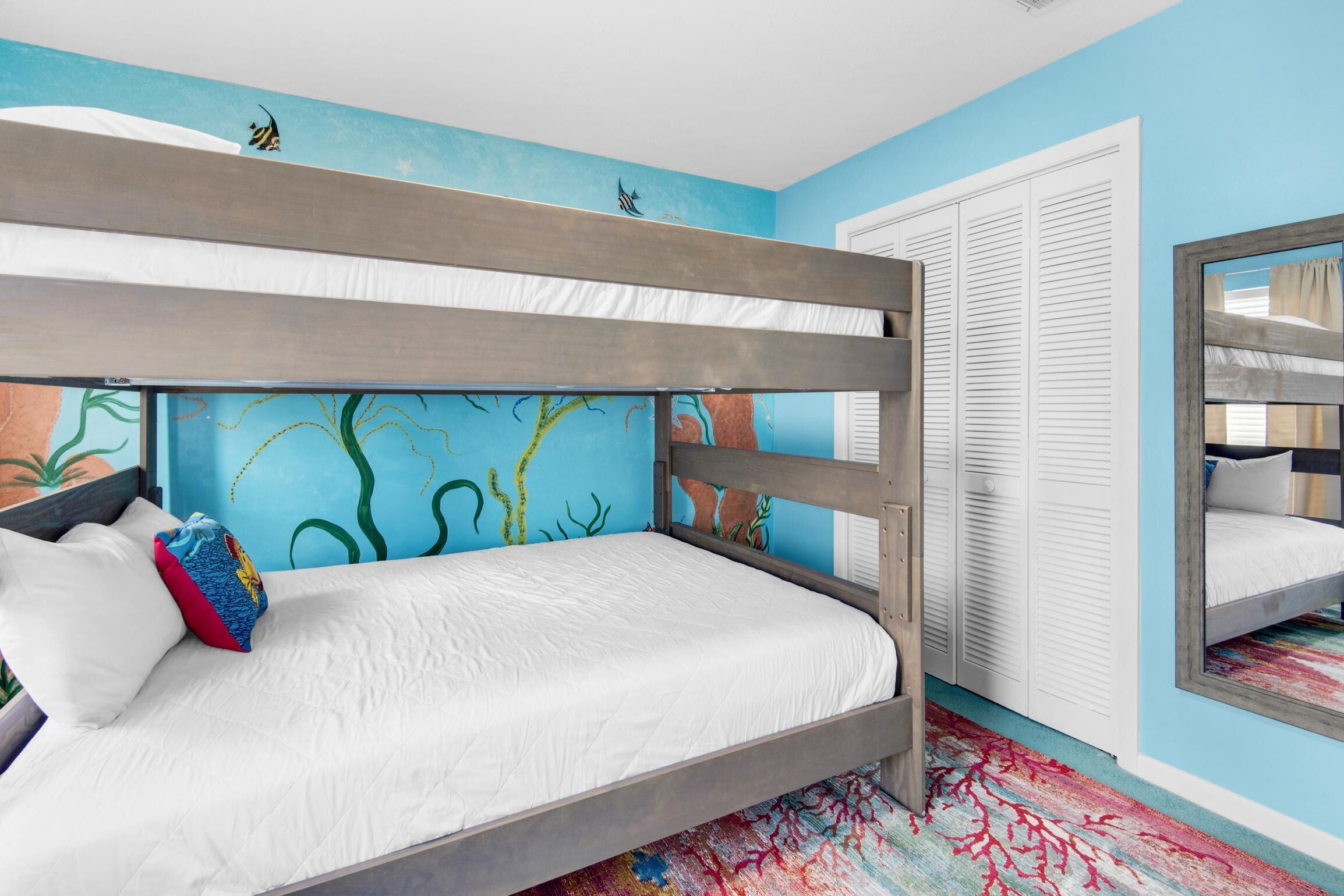 Una cama individual con un colchón blanco y una almohada de colores, un mural con criaturas marinas en la pared, y un armario blanco con puertas correderas.