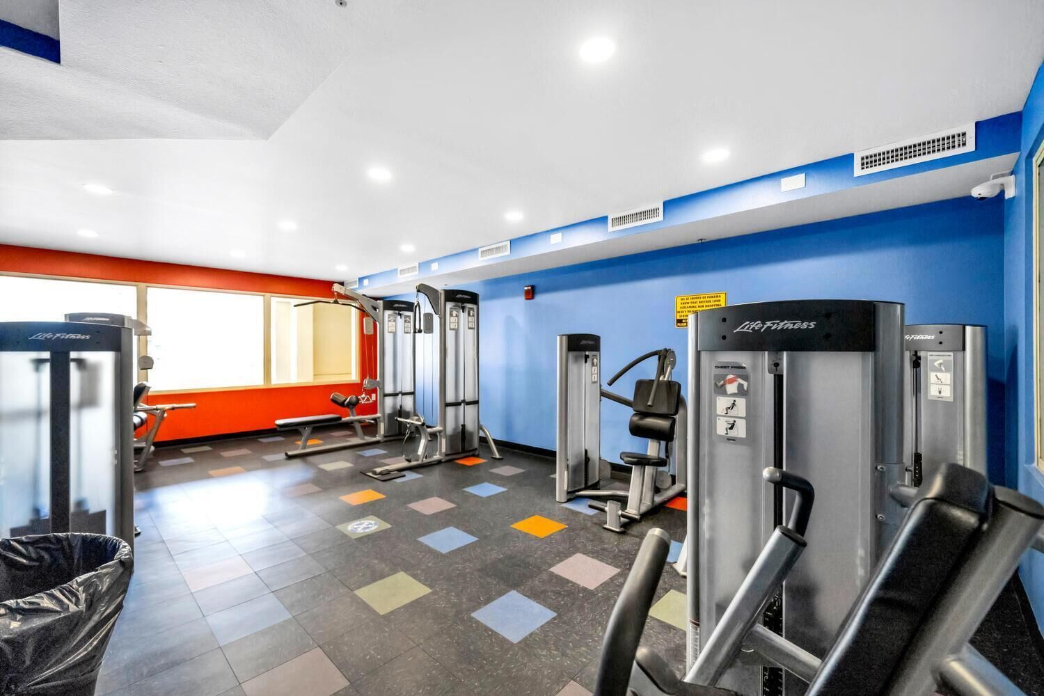 Ein Fitnessstudio mit verschiedenen Trainingsgeräten, einer roten Wand und einem Fenster mit Tageslicht.