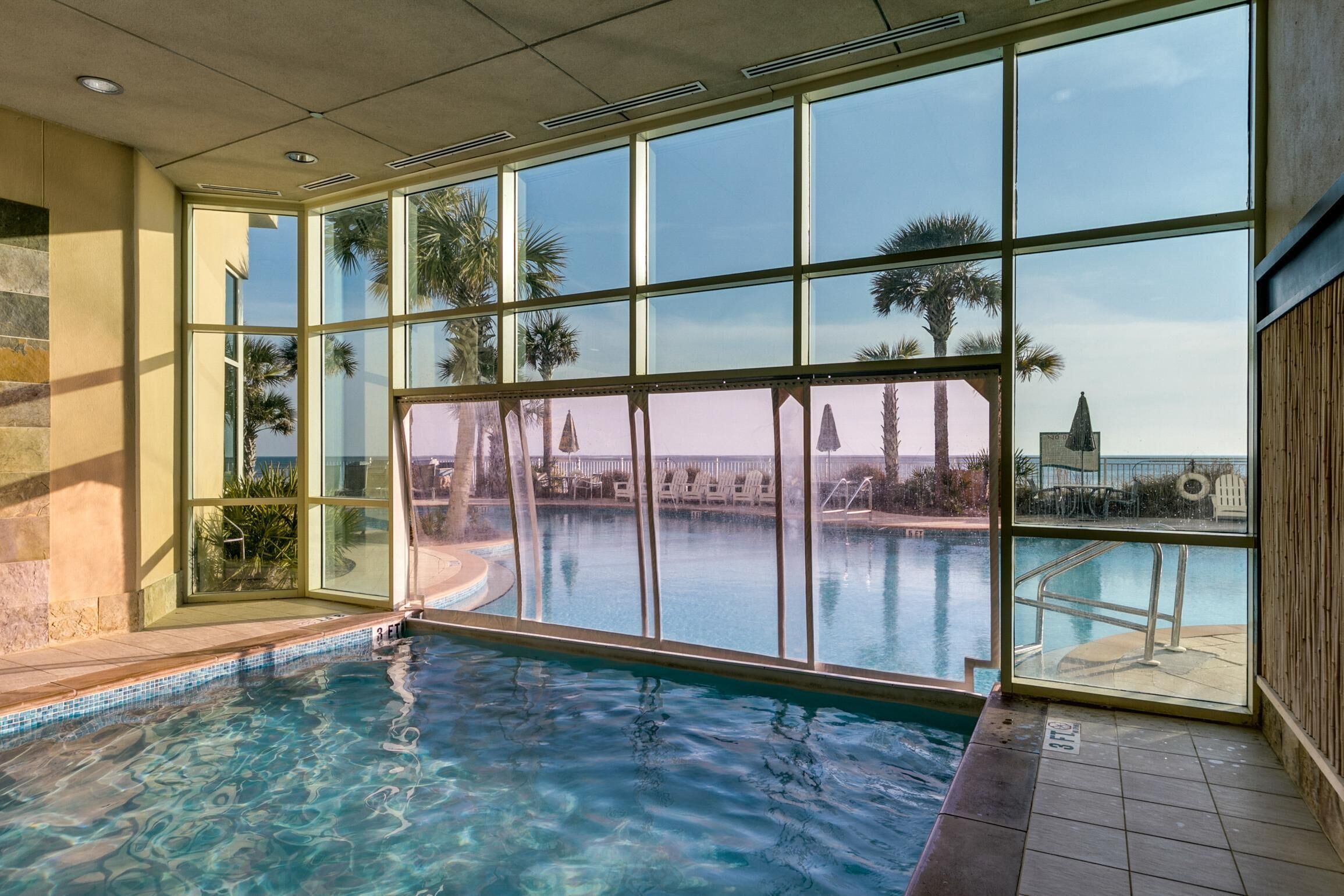Une piscine intérieure avec des parois en verre transparent, un sol carrelé et une vue sur des palmiers et un bâtiment situé au loin.