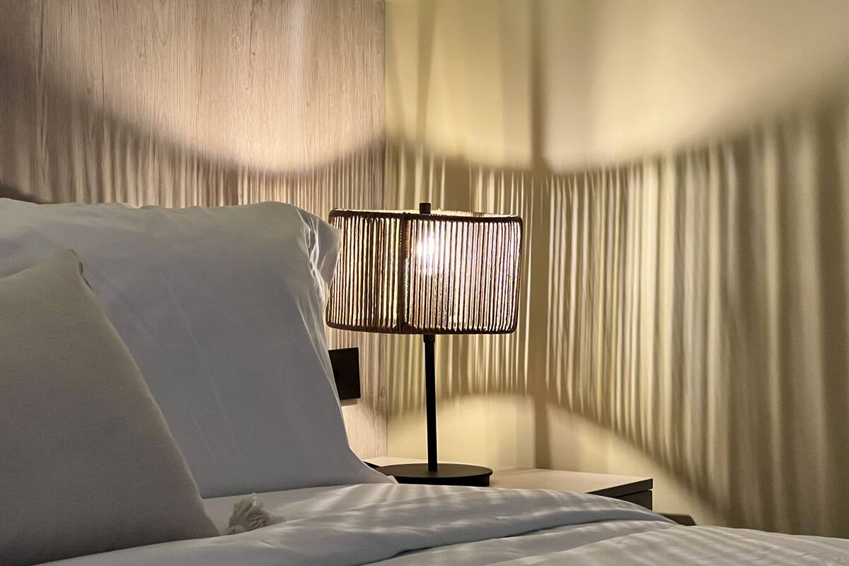 Una cama bien tendida con sábanas y almohadas blancas, una lámpara moderna con pantalla texturizada, y una pared con líneas verticales.