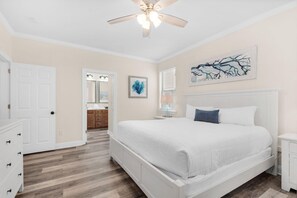 A bedroom with a white bed, a ceiling fan, a door, and a bathroom visible through an open door. - Destin's Siren Song (Destin)