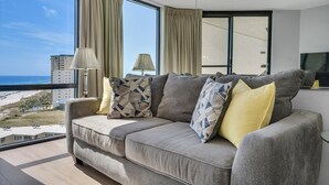 A modern living room with a large window offering a view of the beach and buildings. There is a grey sofa with patterned cushions and two floor lamps. - Sundestin 1415 (Destin)