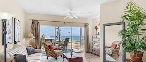 A spacious living room with a large window overlooking the ocean, a ceiling fan, and a dining area with outdoor seating.