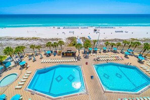 A beachside hotel with two large swimming pools, sun loungers, and palm trees.