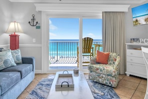 A living room with a sofa, a patterned armchair, a coffee table, and a view of the beach through a sliding door.