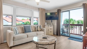 A living room with a sofa, a coffee table, a TV, and a balcony with a view of buildings and trees. - St Thomas Square Villa 1211c Beach Bumz (Panama City Beach)