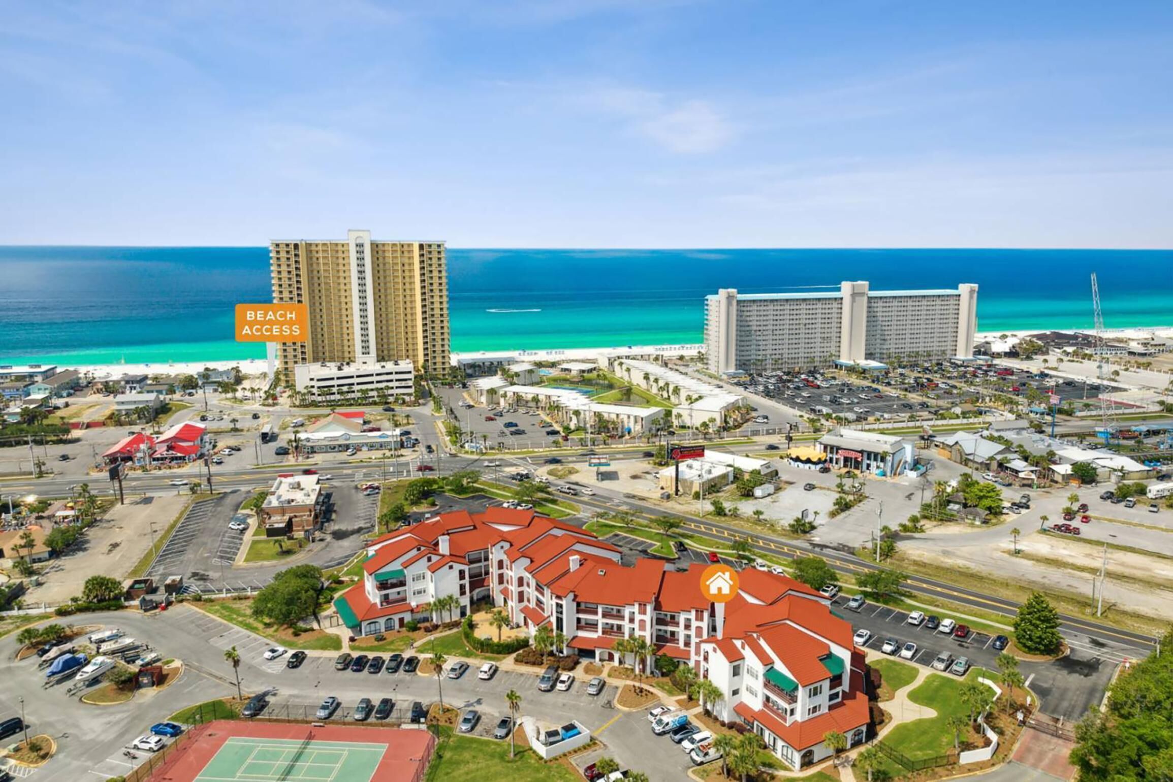 An aerial view of a coastal area with a hotel, a large parking lot, and a beach access sign.