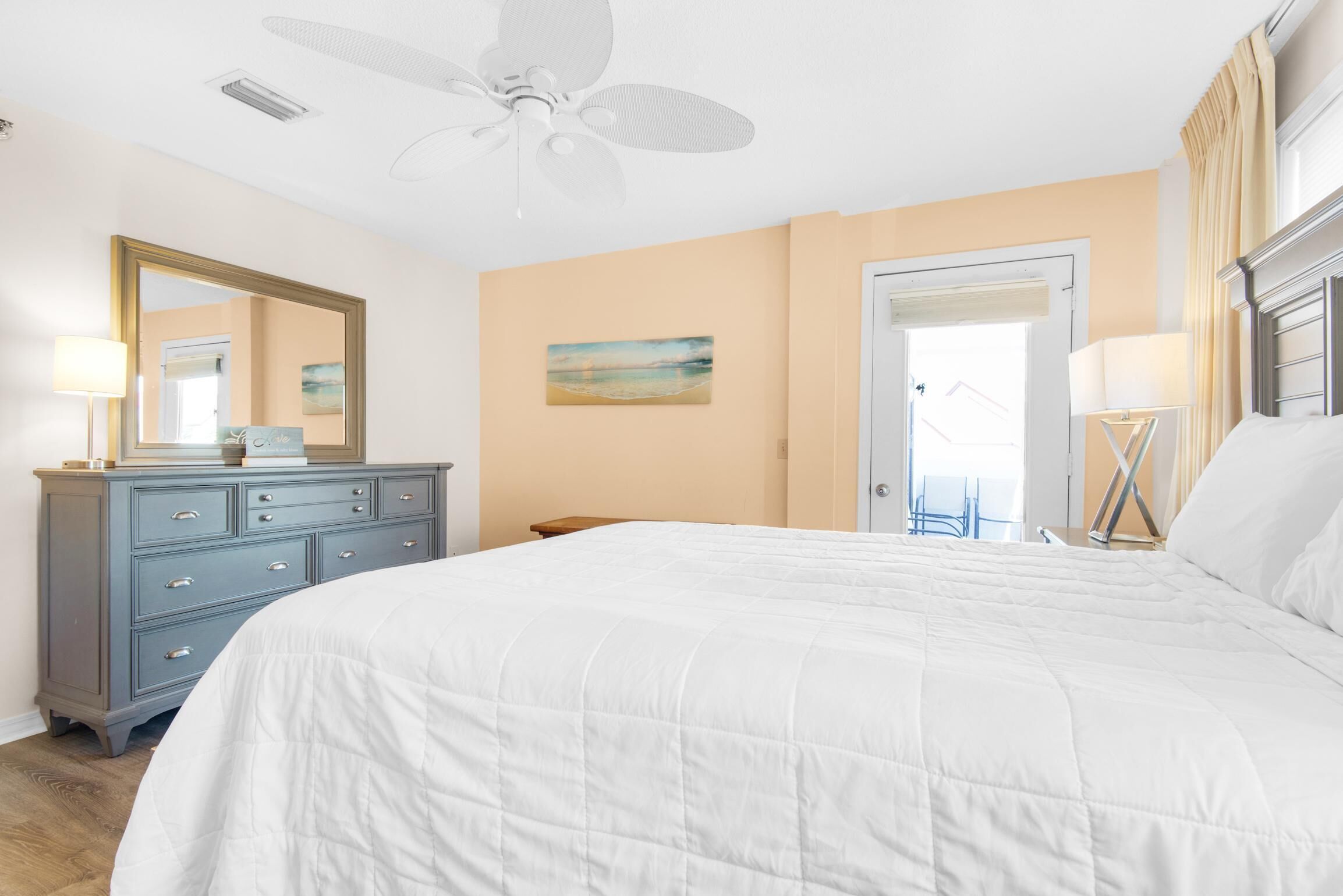 Une chambre avec un grand lit, une commode, un ventilateur de plafond, un tableau accroché au mur et une porte donnant sur une autre pièce.