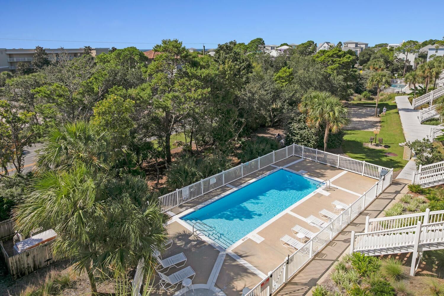 Ein rechteckiger Pool mit weißer Liegefläche und Liegestühlen, umgeben von Bäumen und einer Holzterrasse mit Treppe zu einer erhöhten Ebene.