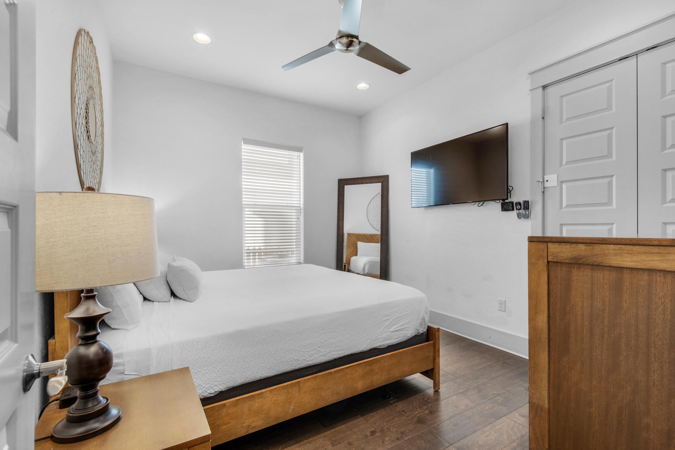 A bedroom with a bed, a ceiling fan, a TV mounted on the wall, and a wooden nightstand with a lamp.