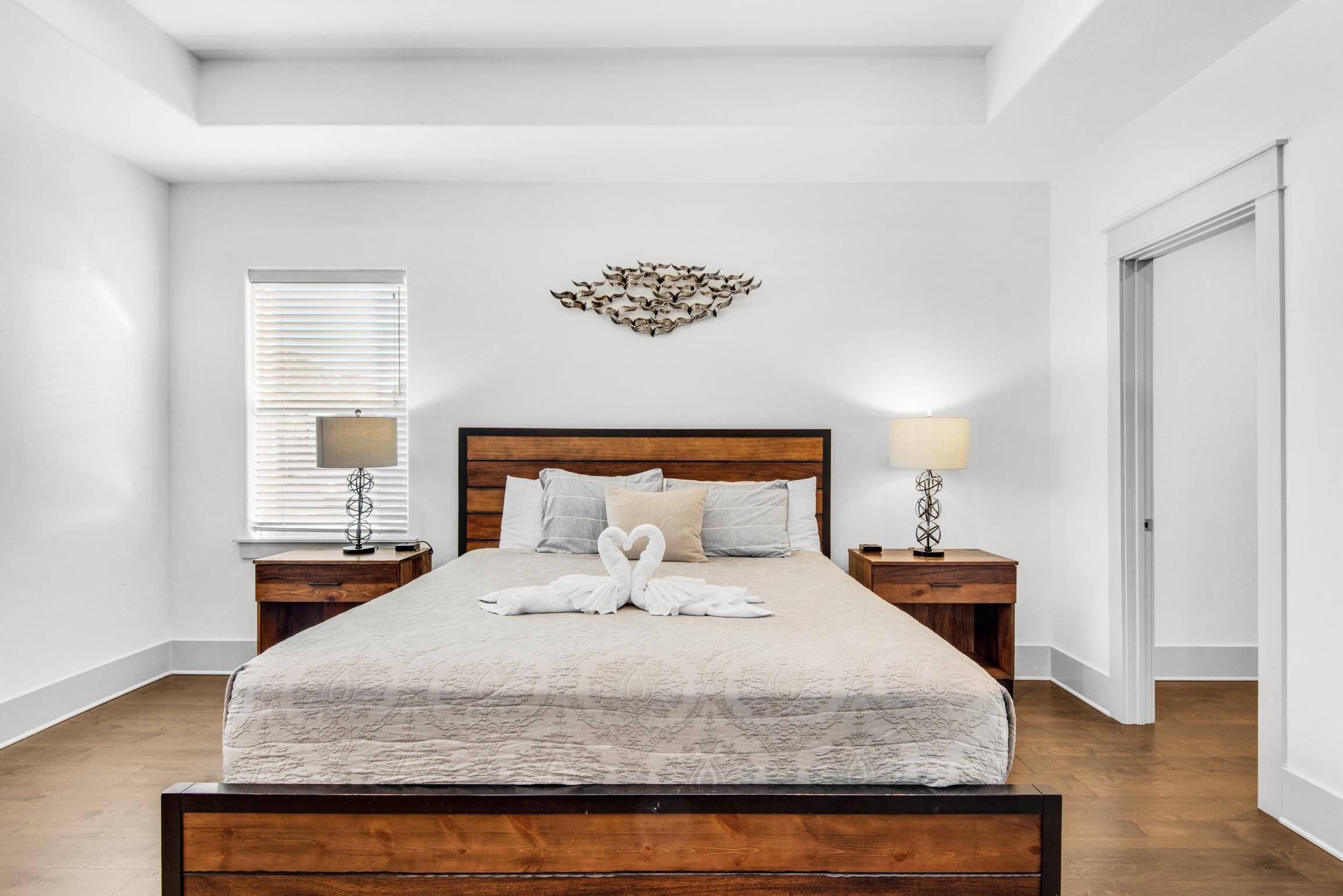 Un dormitorio ordenado con una cama de madera, dos mesitas de noche con lámparas, un adorno en la pared y una ventana con persianas.