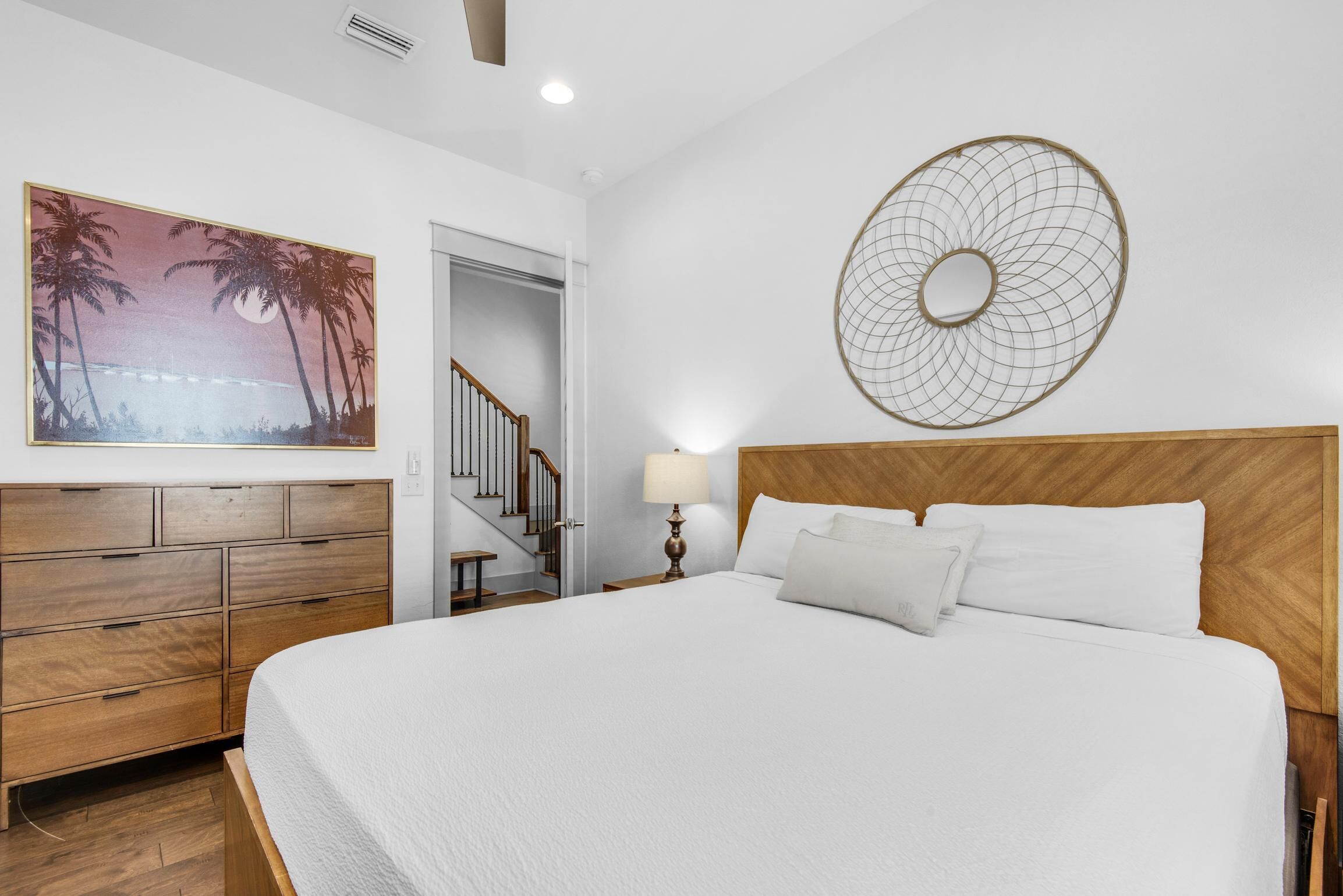 A bedroom with a wooden headboard, a large bed with white bedding, a nightstand with a lamp, a wooden dresser, a wall-mounted circular artwork, and a staircase leading to a closet.