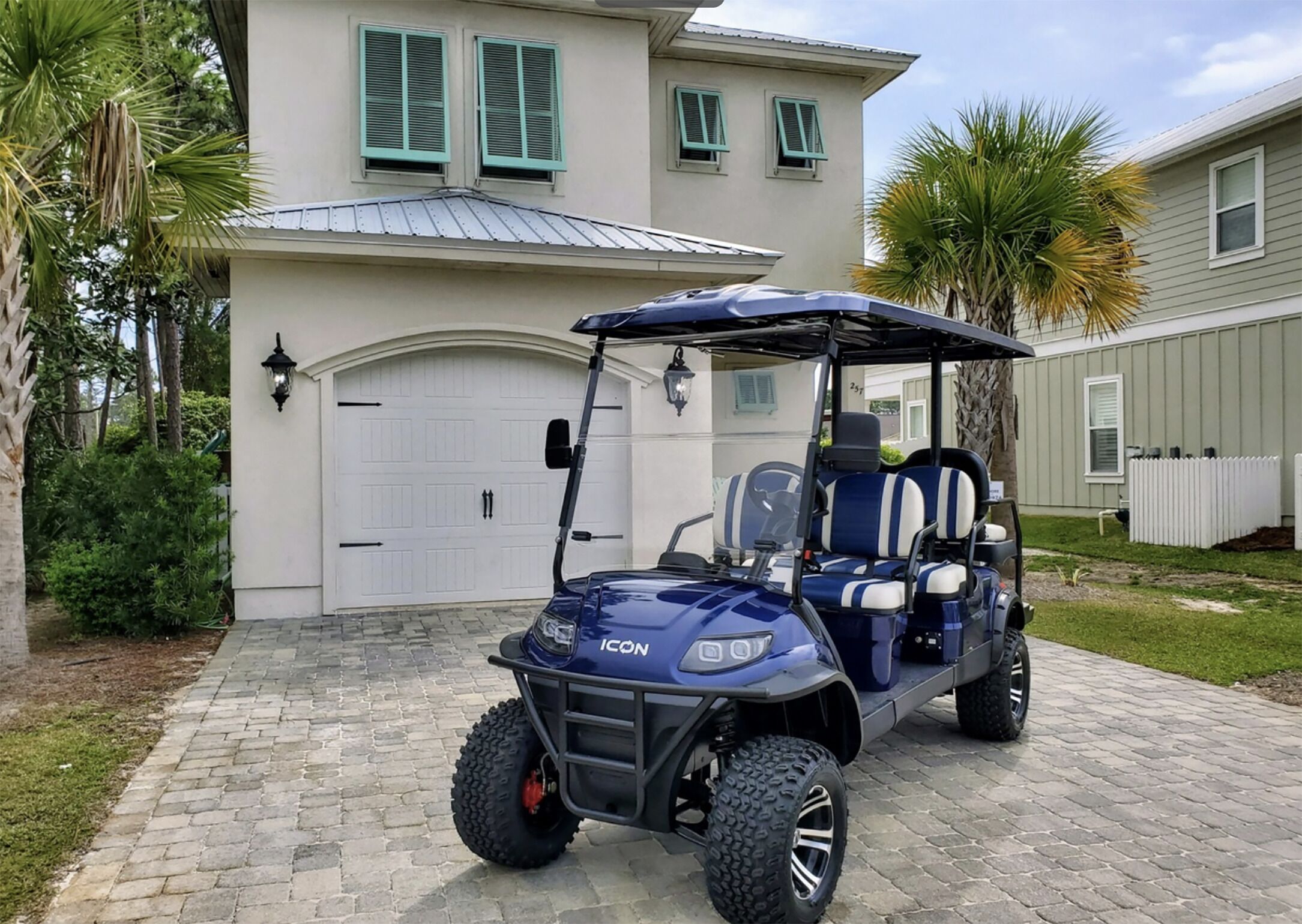 A golf cart parked in front of a house with a garage, a driveway, and a lawn with palm trees.