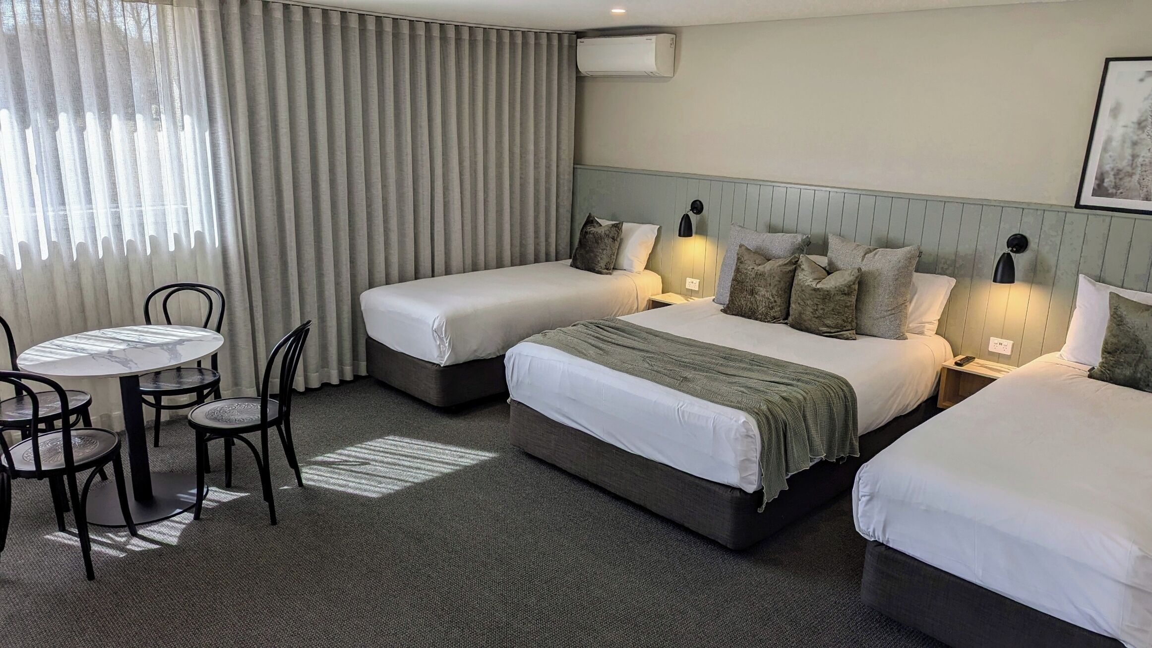 A hotel room with two single beds, a small table with chairs, and a large window with curtains.
