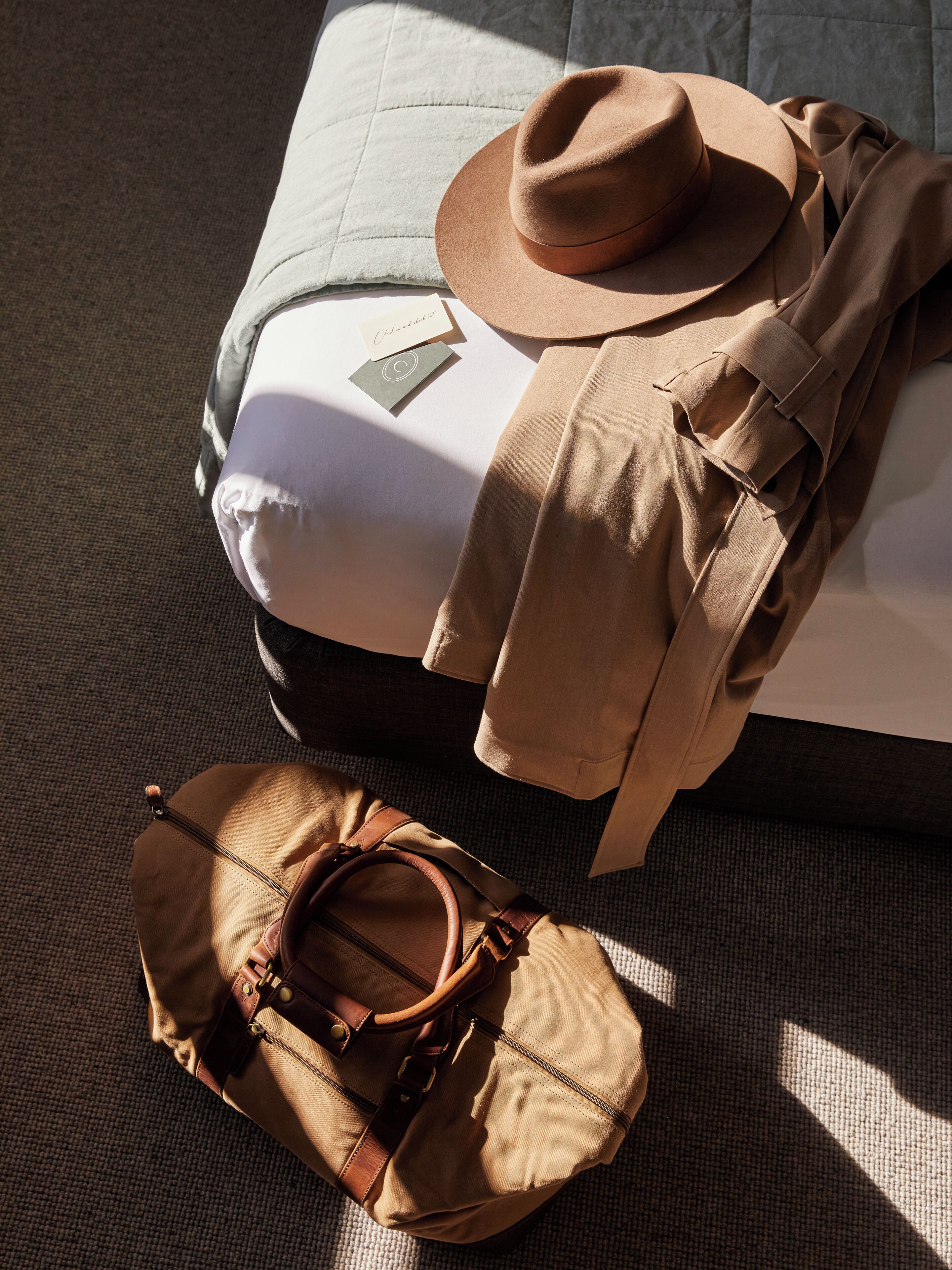 A neatly made bed with a brown hat and a brown bag beside it.