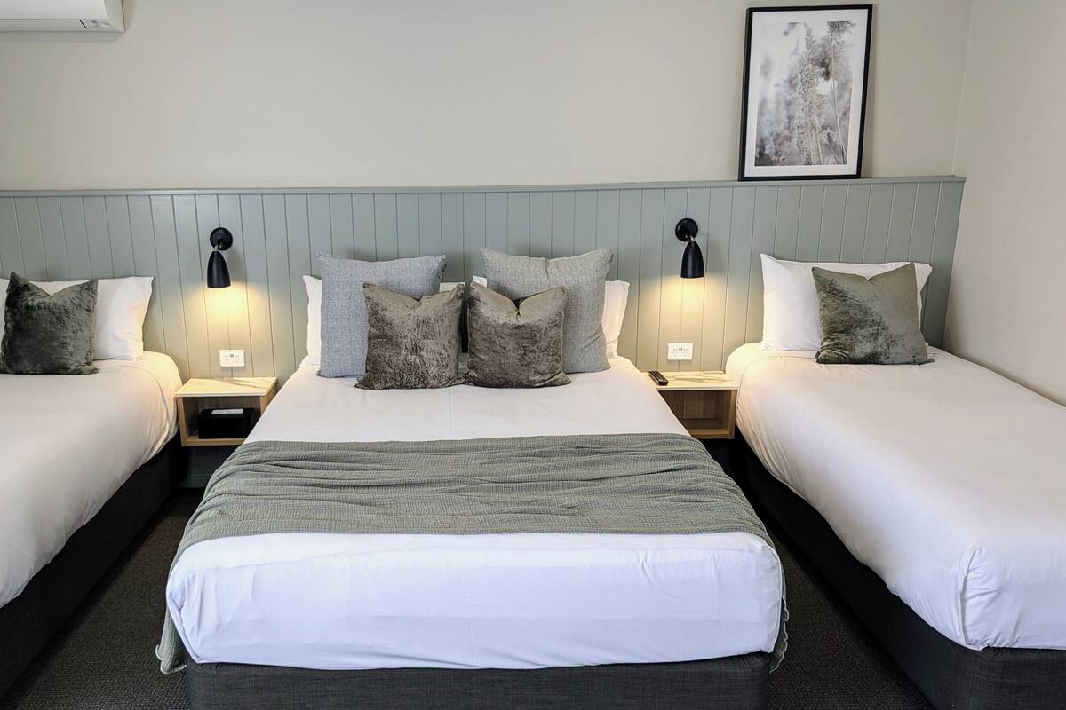 A hotel room with two single beds, a large bed in the center, and a headboard with a light blue panel. There are two bedside tables with lamps, a framed picture on the wall, and a small wooden nightstand under the large bed.