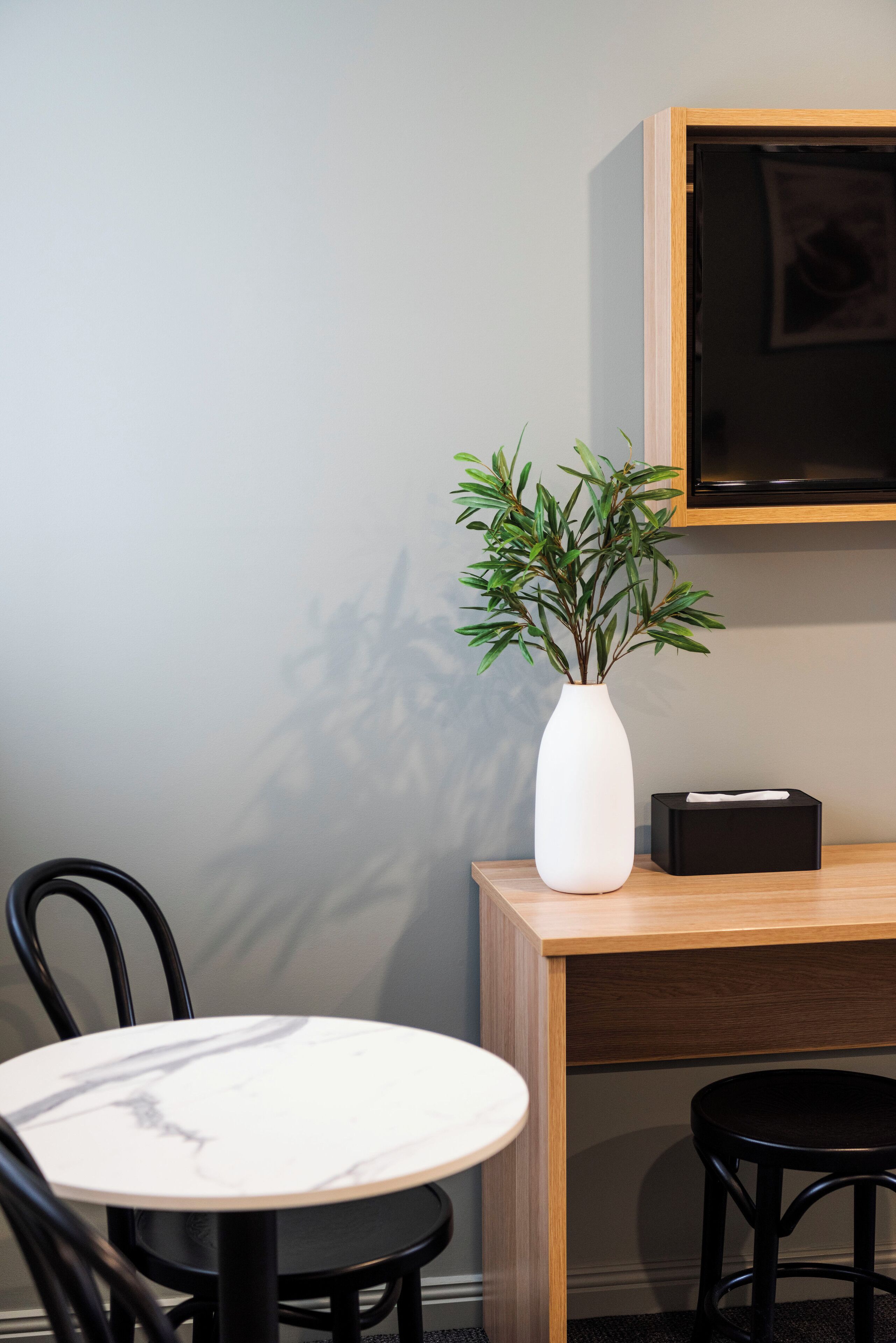 A modern room with a round table, two chairs, a white vase with a plant, a black box, and a mounted television.